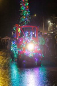 A small red train decorated with colorful Christmas lights and a lit-up tree on top moves down a wet street at night during a festive parade, capturing the warmth of December real estate as people watch from the sidewalks.