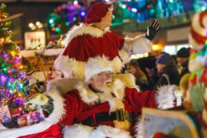 Santa Claus and Mrs. Claus, dressed in festive red outfits, wave and smile atop a holiday float surrounded by colorful lights and a Christmas tree during a nighttime parade, bringing cheer like a real estate update for the winter season.