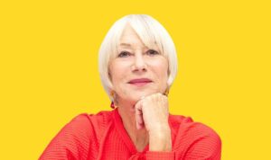 An older woman with short white hair and light skin, wearing a bright red textured top, rests her chin on her hand and smiles softly. Bathed in February light, she sits against a solid yellow background.