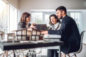 Three people sit at a table in a bright office, smiling and discussing documents. A miniature model of a house sits in the foreground, capturing the spirit of June Joys and new beginnings in this real estate meeting.