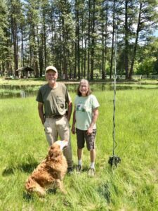 A man and woman stand smiling in a grassy field near a pond and tall pine trees, with a golden retriever at their feet. Nearby, a tall pole with equipment—perhaps for community lights—adds interest to the peaceful scene.