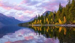 A calm lake reflects a vibrant forest with autumn-colored trees and distant mountains under a colorful, partly cloudy sky at sunset—capturing the spirit of August Reflections.