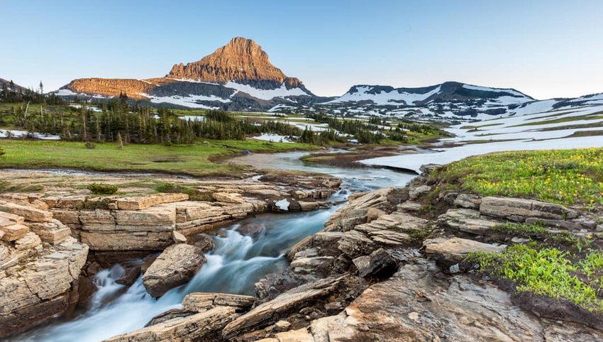 A clear mountain stream flows over rocky terrain with patches of green grass and wildflowers, celebrating June Joys as it leads to a snow-dotted landscape and a prominent peak under a clear blue sky.