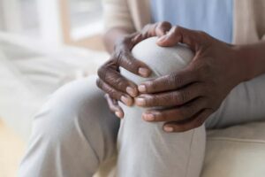 A person sitting and holding their knee with both hands, suggesting pain or discomfort in the knee area. The image focuses on the hands and knee, with a softly blurred background evoking the gentle renewal of spring.