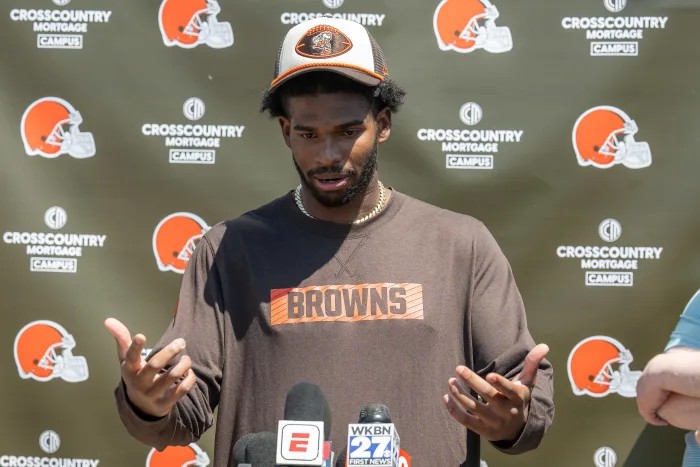 A Cleveland Browns football player speaks at a press conference, standing in front of a Browns-branded backdrop with microphones in front of him, wearing a Browns shirt and cap, sharing his August Reflections ahead of the Season of Action.