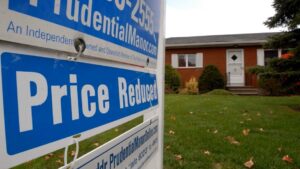 A Price Reduced real estate sign stands in front of a single-story brick house with a green lawn and shrubs, inviting buyers to enjoy new beginnings and seize June joys with this attractive home for sale at a lower price.