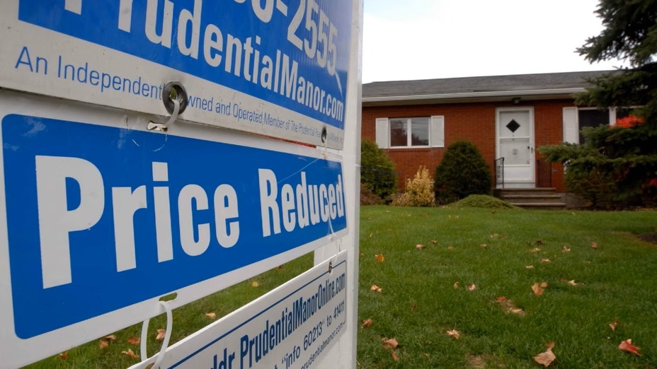 A Price Reduced real estate sign stands in front of a single-story brick house with a green lawn and shrubs, inviting buyers to enjoy new beginnings and seize June joys with this attractive home for sale at a lower price.