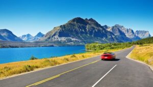 A red car drives on a winding road next to a large blue lake, with tall, rugged mountains and a clear blue sky in the background, capturing the steady momentum of travel beneath the crisp February light.