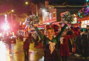 A smiling cheerleader in a sparkly outfit and large white bow raises pom-poms during a festive nighttime parade, capturing the winter glow as colorful lights shine behind other performers.