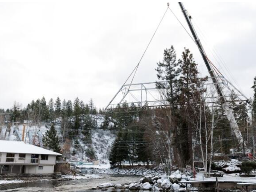 A large crane lifts a metal bridge frame over a snowy riverside landscape with trees, a house, power lines, and rocky banks—capturing the Winter Pause before Spring Success transforms the scene.