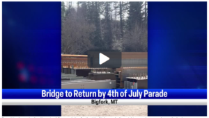 Video screenshot with construction materials and beams stacked outdoors, set against a backdrop of trees. A news banner reads: Bridge to Return by 4th of July Parade, Bigfork, MT. Spring Into the Season with this exciting community update! A play button is centered on the image.