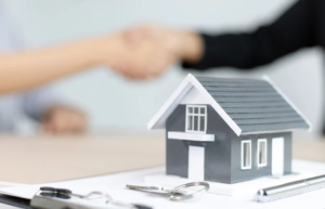 A small model house sits on a desk with keys and a clipboard in front, while two people in the background shake hands, marking a real estate agreement—a perfect scene for the spring season of home buying.