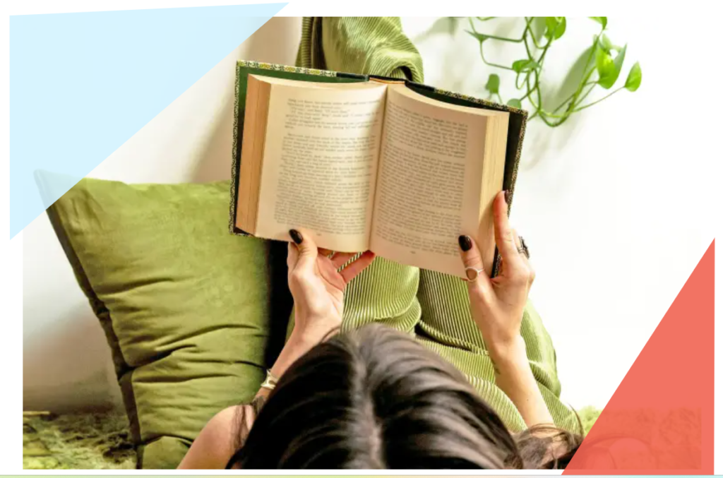 A person with dark hair lies on green cushions, holding an open book above their face. A green plant hangs in the background, while pastel blue and red geometric shapes frame the corners—capturing a quiet August Reflections moment.