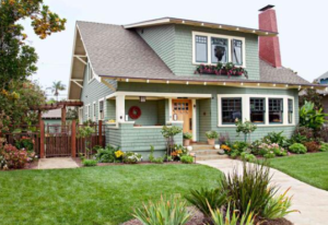 A two-story house with green shingles, a red brick chimney, and white trim, surrounded by a well-kept lawn and colorful flowers—perfect for when love is in the air and the market heating up. A wooden fence with an arbor graces the left side.