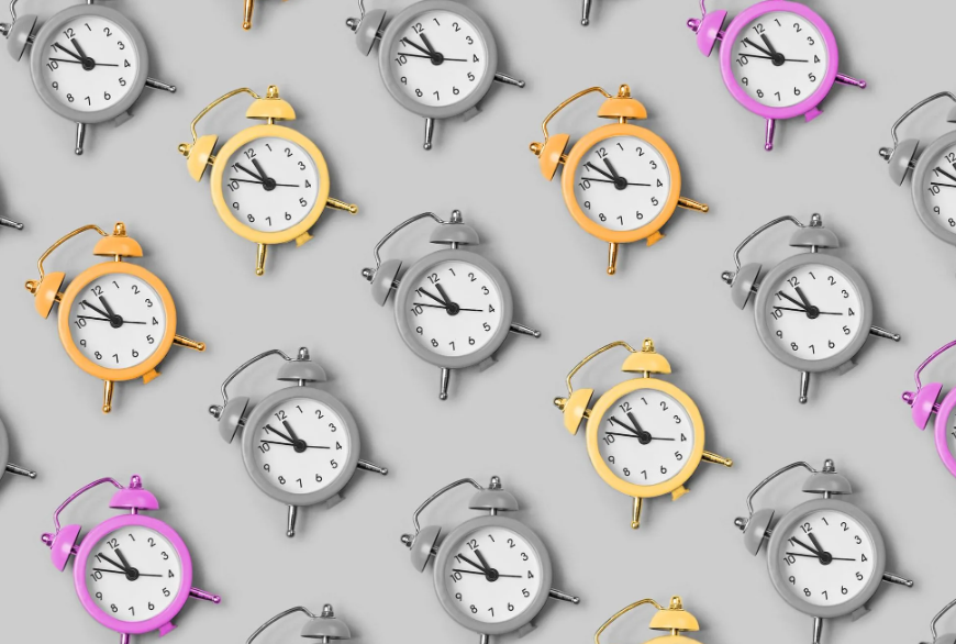 A seamless pattern of vintage alarm clocks in yellow, gray, and purple, set against a light gray background. Reminiscent of the Ides of March, the clocks display various times—suggesting new opportunities as spring arrives.