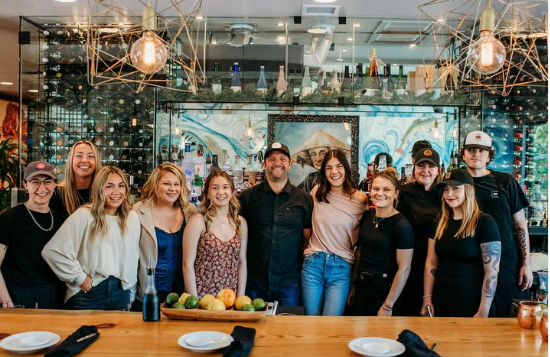 A group of eleven people, smiling and standing together behind a wooden counter in a modern bar or restaurant, with bottles and decorative lights in the background. A tray full of fruit sits on the counter in front of them.