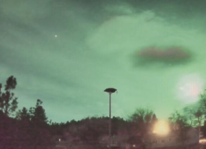A green-tinted night sky with a bright glowing object near the horizon, silhouetted trees and houses, and a single streetlight in the foreground capture the cozy season’s serene community spirit.