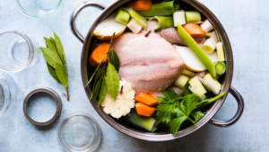 A stainless steel pot contains a whole raw chicken, carrots, celery, leeks, garlic, and bay leaves—perfect for a cozy season meal. Several empty glass jars and lids are arranged nearby on a light surface.
