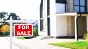 A red and white For Sale sign is posted in front of a modern house with large windows and a well-kept lawn, offering buyers a great opportunity in the Flathead Valley spring market.