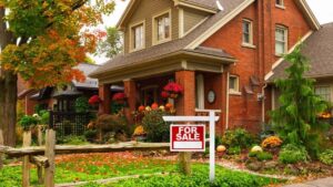 A red-brick house with a well-kept garden decorated for autumn, featuring pumpkins and flowers, stands behind a wooden fence and a For Sale sign, surrounded by trees with colorful fall leaves.