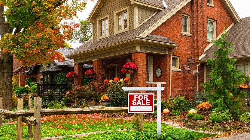 A red-brick house with a well-kept garden decorated for autumn, featuring pumpkins and flowers, stands behind a wooden fence and a For Sale sign, surrounded by trees with colorful fall leaves.