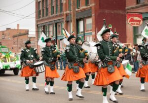 A marching band in green jackets and orange kilts plays bagpipes and drums during the Flathead Valley parade. Shamrock decorations and a float are visible, with people watching from the sidewalk in front of brick buildings.
