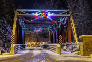 A snow-covered bridge at night is decorated with colorful Christmas lights, garlands, and a neon Merry Christmas sign, capturing the intention of new beginnings. Snowy trees and a warmly lit house glow in the background.