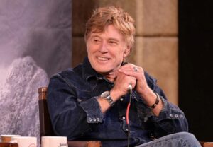 An older man with light brown hair, wearing a denim shirt and jeans, sits smiling on stage while holding a microphone. Seated before a mountain backdrop, he radiates intention as he speaks, a coffee mug beside him hinting at new beginnings.