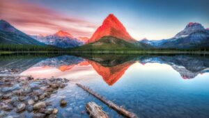 A serene lake reflects a red-tinged mountain at sunrise, surrounded by forest and other peaks beneath a blue sky. Along the shore, rocks and driftwood hint at Winter Pause giving way to the vibrant promise of Spring Success.