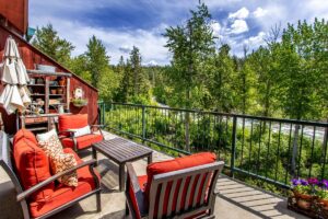 A cozy outdoor patio with red cushioned chairs and a table overlooks a lush, green forest and river under a partly cloudy sky. Potted plants and decorative items celebrate June joys, adding charm to this inviting space.