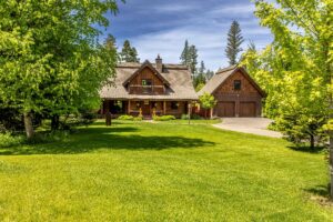 A rustic log cabin with a covered front porch and attached garage sits amid lush green grass, trees, and landscaping under a blue sky with scattered clouds—perfect for carefree Lake Days or exploring new Life Listings.