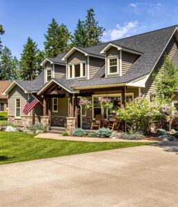 A two-story suburban house with gray siding, stone accents, and a covered front porch. Perfect for those beginning the sellers process, it features rocking chairs, hanging flower baskets, an American flag, and a landscaped yard with green grass and shrubs.