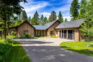 Modern single-story house with wood siding, black trim, and a metal roof, featuring a double garage with glass-panel doors—perfect for July Journeys or relaxing Lake Days—surrounded by green lawn, trees, and a clear blue sky.