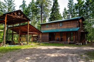 A rustic wooden house with a green metal roof sits in a forested area, next to an open-sided wooden shelter. Tall pine trees surround the buildings, and outdoor chairs are visible on the porch, evoking local folklore and community lights gatherings.