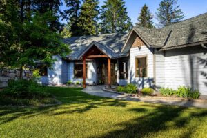 Single-story house with light blue siding, large wooden front porch, and brown trim, surrounded by green grass and a landscaped garden. Use our Mortgage Calculator to estimate monthly payments for this serene home under a clear sky and tall trees.