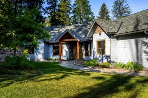 A single-story house with light blue siding, a dark gray roof, and wooden entrance beams stands amid green grass, plants, and tall trees in a sunny yard—radiating intention and the promise of new beginnings.