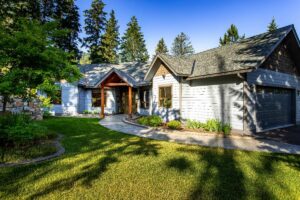 Single-story house with gray siding, a gabled roof, and attached garage, surrounded by green lawn and tall trees under a clear blue sky. Sunlight casts shadows, evoking August Reflections on the peaceful scene.
