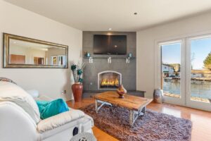 A cozy living room with a fireplace under a wall-mounted TV, a wooden coffee table on a shaggy rug, and a white sofa with teal accents—perfect for Lake Days, with large windows overlooking a serene waterfront view.
