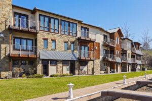 Modern three-story townhouse building with stone and wood exterior, multiple balconies with metal railings, large windows, and a green lawn in front—perfect for Blooming Homes—beside a wooden walkway under a clear blue sky.