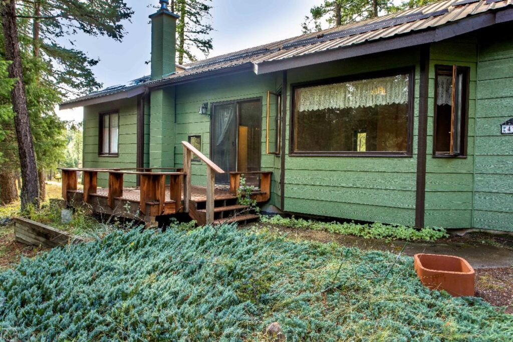 A green house with large windows, a wooden porch with railings, and steps leading to the entrance, surrounded by trees and plants in vibrant autumn colors, under a partly cloudy sky.