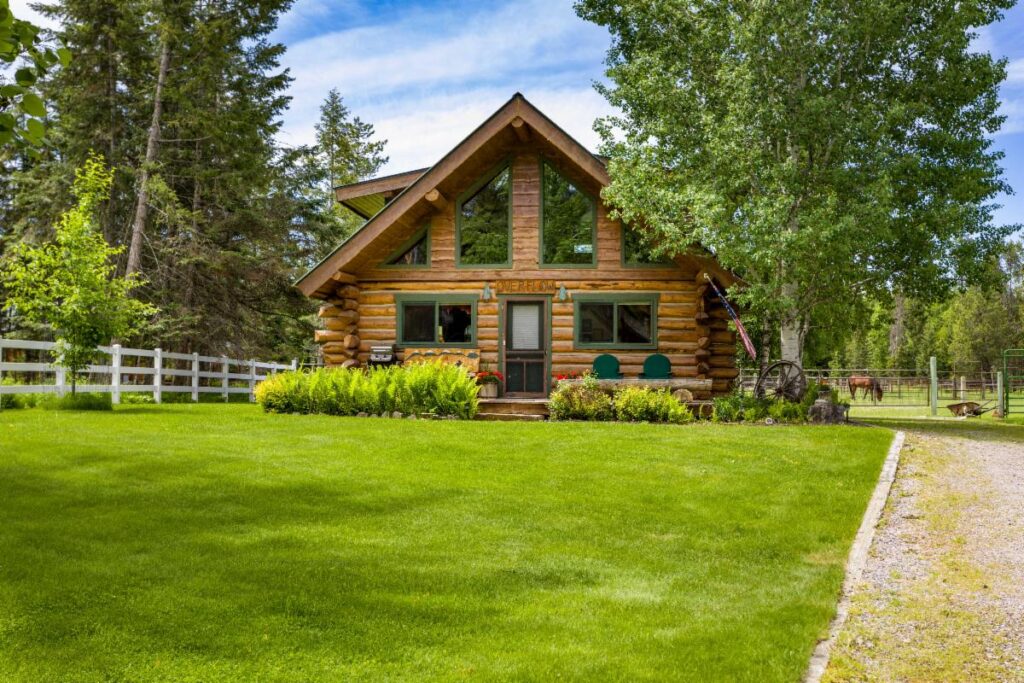A cozy log cabin with large windows sits on a lush green lawn, surrounded by trees and a white fence, under a blue sky with clouds—a perfect spot for August Reflections in this tranquil Season of Action.