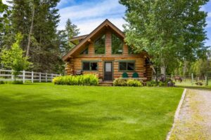A cozy log cabin with large triangular windows sits on a lush green lawn, surrounded by trees and a white fence, basking in the soft Winter Glow under a blue sky with scattered clouds.