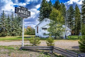 A vintage sign reading LaSalle ANGEL THEATER stands in front of a white, weathered building, its community lights glowing among tall pine trees and a grassy yard under a partly cloudy sky.