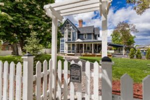 A charming blue and white Victorian-style house with a covered porch, viewed through a white picket fence and arbor, is surrounded by green lawn and trees—a perfect scene for cool crisp days during the changing season.
