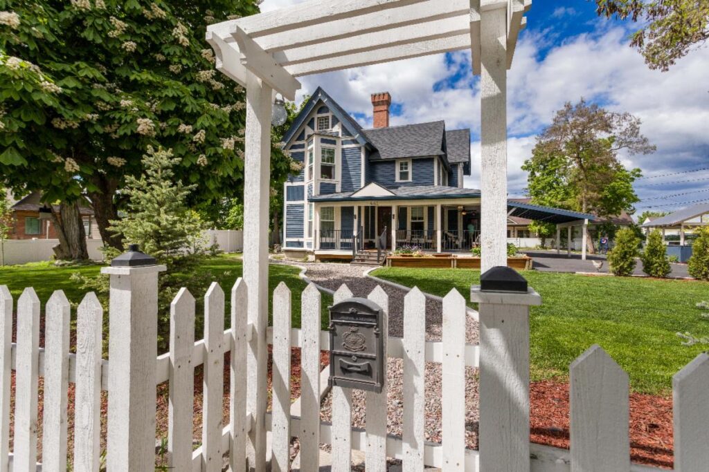 A charming blue and white Victorian-style house with a front porch, viewed through a white picket fence and arbor, surrounded by lush green lawn, colorful autumn trees, and a partly cloudy sky.