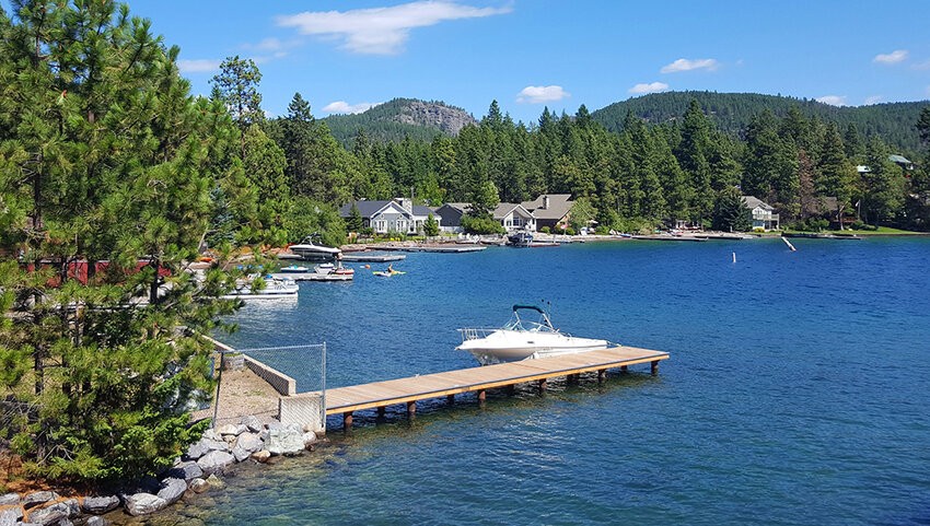 A peaceful lakeside scene with a wooden dock holding a white boat, several houses among pine trees, blooming homes along the shore, and forested hills in the background under a blue sky.