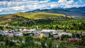A scenic view of a small town nestled among green hills and dense trees, with a river in the foreground and snow-capped mountains beyond, evokes the warmth of Merry Christmas under a partly cloudy sky.