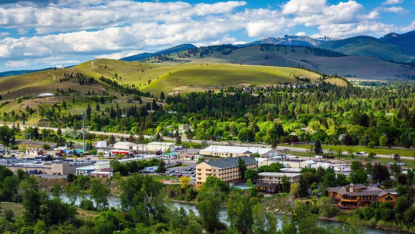 A scenic view of a small town nestled among green hills and dense trees, with a river in the foreground and snow-capped mountains beyond, evokes the warmth of Merry Christmas under a partly cloudy sky.