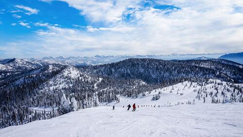 Skiers descend a snowy mountain slope surrounded by snow-covered trees and distant mountain ranges under a partly cloudy blue sky, embracing the crisp air of fall in this October adventure.