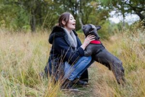 A woman kneels in tall grass outdoors, smiling and holding a gray French Bulldog in a red harness. Surrounded by trees and greenery, they savor Lake Days together—capturing the joy of July Journeys in nature.